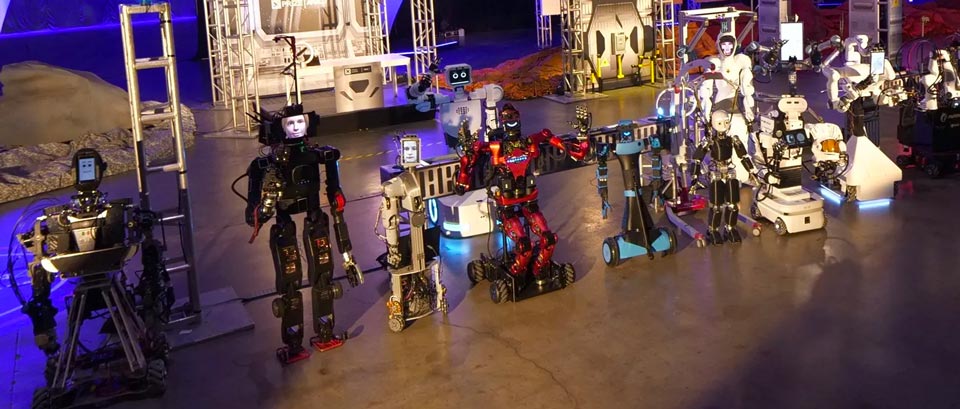 Дюжина різних роботів, що вишикувалися поруч один з одним на арені змагань